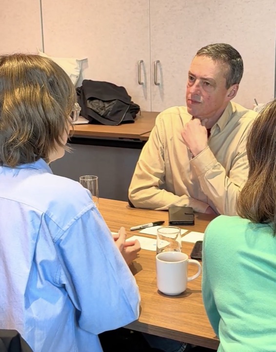 Three participants in conversation at a table with notebooks and coffee cups.