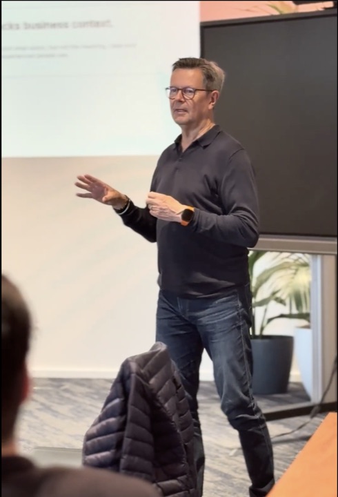 A speaker gesturing beside a projected slide and a large display.